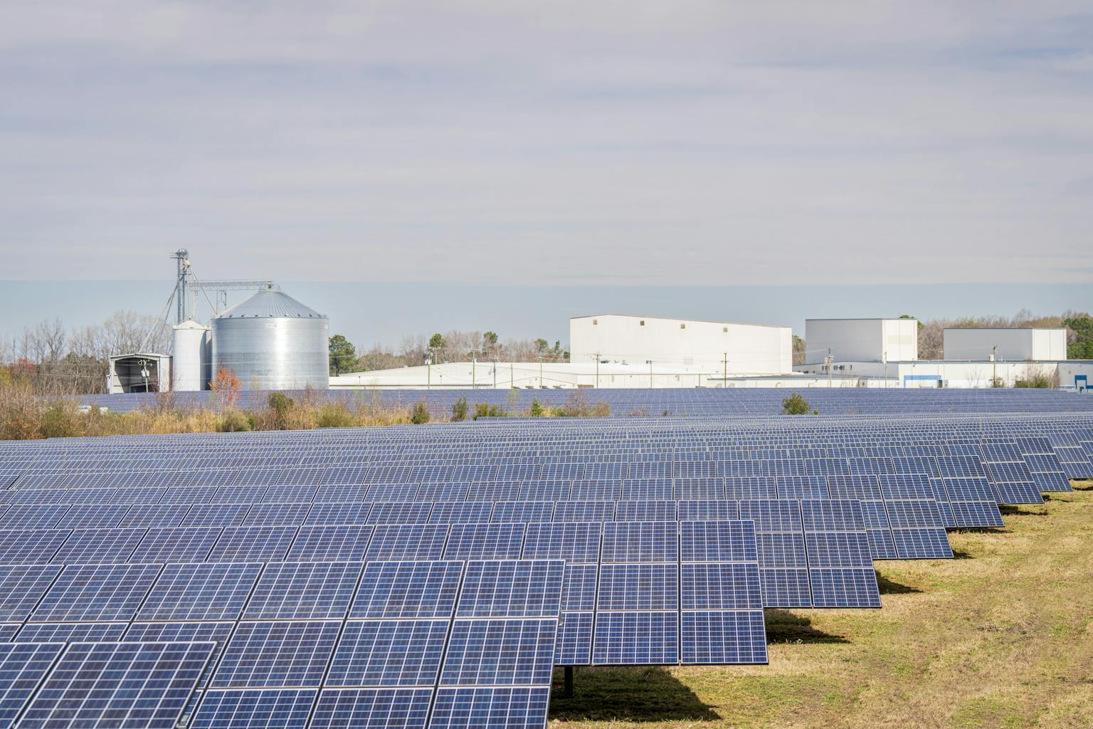 A large solar panel field with warehouses and silos in the background under a clear sky.
