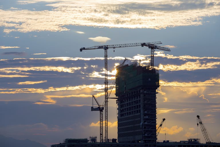 Silhouette of cranes and a building under construction against a vivid sunset sky.